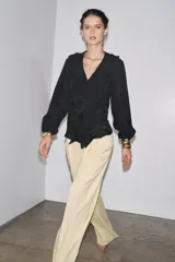Camisa color rosa pálido confeccionada en tejido ligero con lyocell. Presenta escote en V, cierre frontal con botones, mangas largas abullonadas con puños elásticos y detalle de volantes en el frente.