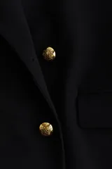 Blazer negro de corte recto, doble botonadura con botones dorados, solapas de pico, hombreras, bolsillos delanteros ribeteados con solapa, botones decorativos en los puños y abertura trasera.