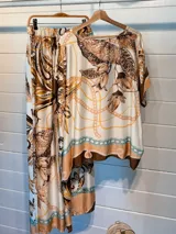 Pantalón ancho de satén con cintura elástica y estampado de motivos florales y cadenas en tonos marrones, beige y dorado sobre fondo blanco. Presenta un borde inferior con detalles en color turquesa.