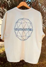Remera color crema de corte amplio con cuello redondo y mangas cortas. Estampa circular en el frente con diseño geométrico y la palabra "Vancouver" en color azul.