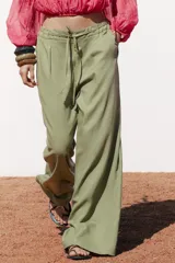 Pantalón ancho color verde oliva confeccionado en lino. Tiro alto con cintura elástica ajustable con cordón y pinzas delanteras. Presenta bolsillos laterales y traseros. Incluye cinturón de la misma tela con apliques metálicos.