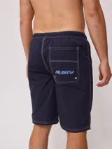 Short de baño azul marino con cintura elástica ajustable con cordón, bolsillos laterales y logo de la marca Rusty estampado en azul en la pierna izquierda.