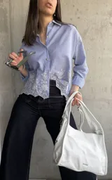 Camisa celeste a rayas verticales blancas, con cuello clásico, cierre frontal con botones y mangas largas. Presenta detalle de bordado calado con motivos florales en el ruedo.