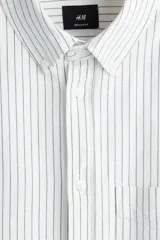 Camisa de manga larga color blanco roto con finas rayas verticales en tono oscuro. Presenta cuello americano, bolsillo superior abierto y corte estándar.