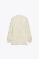 Blusa color crudo confeccionada en viscosa, con cuello redondo, escote en pico, mangas largas con puños elásticos, volantes frontales y detalles de encaje.