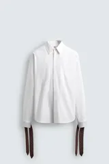 Camisa blanca de algodón popelín, corte relajado, con cuello solapa y pechera frontal plisada. Las mangas terminan en puños ajustables con lazos largos estampados en color marrón con patrón circular.