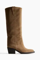 Botas de caña alta estilo cowboy, color marrón claro (camel/taupe), con acabado de gamuza o nobuck. Presentan costuras decorativas en la caña, punta de almendra y taco ancho y bajo de color oscuro.