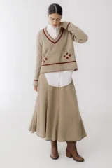 Sweater beige con cuello en V y mangas largas con diseño trenzado. Tiene dos bolsillos frontales con bordados de tréboles rojos y ribetes rojos en cuello, puños y ruedo.