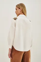 Camisa blanca de algodón con cuello clásico, mangas largas abullonadas con puños anchos y cierre frontal de botones. Presenta aplicaciones bordadas en dorado con detalles de animal print en los hombros.