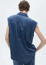 Camisa azul sin mangas de tejido fluido y satinado, con cuello camisero, cierre frontal con botones y bolsillo de parche en el pecho.