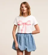 Remera blanca de algodón con cuello redondo y mangas cortas. Presenta una estampa de una moña rosa en el frente.