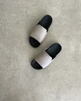 Sandalias chatas color beige con base negra y detalle de bordado blanco en la tira superior.