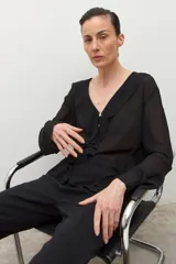 Blusa negra de manga larga, confeccionada en tela liviana y semitransparente, con escote en V adornado con volados y un botón frontal.