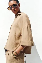 Camisa color beige de cuello solapa con escote pico y manga corta ancha acabada en vuelta con trabilla y botón. Cierre frontal con botones.