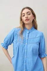 Camisa celeste de gasa de algodón con textura cuadrillé, cuello con volados, mangas abullonadas con puño elástico e inserciones de encaje lineal en el frente.