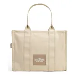 Bolso tote grande de lona color beige con asas de mano y estampado frontal con el texto "THE TOTE BAG MARC JACOBS" en blanco.