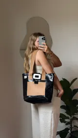 Bolso tote de ecocuero marrón oscuro con textura cocodrilo y un parche frontal de cuero liso marrón claro. Posee asas de hombro de cuero marrón y un broche decorativo blanco en el frente.