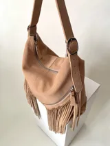 Bolso tipo hobo color tostado, hecho de cuero con flecos largos en el frente y dos bolsillos con cierre. Tiene una manija regulable con hebillas metálicas.