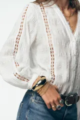 Blusa blanca de manga larga con escote en pico, detalle de tejido con frunces, entredoses y plumeti. Cierre frontal con botones.