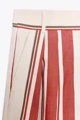 Pantalón de tiro medio con estampado de rayas verticales negras sobre fondo blanco. Tiene trabillas en la cintura, bolsillos laterales, pinzas delanteras y cierre frontal con cremallera, botón y ganchos metálicos.