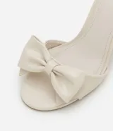 Sandalias de tacón alto color crema, con punta redonda abierta, tira fina con hebilla en el tobillo y maxi lazo decorativo en la parte delantera.