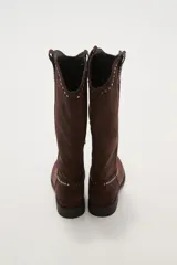 Botas texanas negras de caña media, confeccionadas en gamuza. Presentan un diseño de estilo western con detalles de tachas metálicas plateadas decorando el borde superior y el contorno del empeine. Tienen un taco bajo y punta ligeramente afilada.