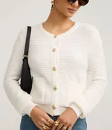 Cardigan de punto blanco, con cuello redondo, manga larga y cierre frontal de botones dorados. Posee dos bolsillos cuadrados en la parte delantera inferior.