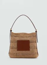 Bolso tipo tote de paja trenzada color beige con aplique rectangular de cuero marrón en el frente. Tiene forro interior, cierre con botón magnético y asa de hombro ajustable.