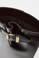 Bota alta de cuero color marrón oscuro, con caña alta, punta redonda y hebilla decorativa en la parte superior.