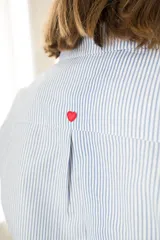 Camisa celeste a rayas verticales blancas, de corte clásico con cuello camisero, mangas largas y cierre frontal con botones rojos. Presenta costuras en contraste y un corazón bordado en la espalda.