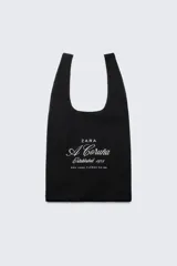 Bolso tipo shopper negro con asas cortas. Estampado frontal con el logo de Zara, la leyenda "A Coruña Established 1975" y la dirección "Rúa Juan Flórez 64-66" en color blanco.