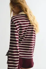 Remera tipo polo de manga larga con corte holgado, a rayas horizontales color borgoña y crema. Tiene cuello clásico color crema, tapeta con botones y hombros caídos.