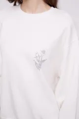 Buzo blanco de corte holgado con cuello redondo y mangas largas. Presenta un pequeño estampado de una flor en el centro del pecho.