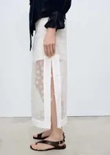 Falda midi blanca con bordado de flores amarillas, diseño semitransparente, tiro medio, corte recto, abertura lateral y cierre de cremallera lateral.