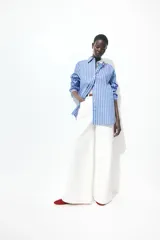 Camisa oversized de popelina de algodón a rayas verticales celestes, blancas y rojas. Tiene cuello clásico, cierre frontal con botones, hombros caídos y mangas largas con puños abotonados. El bajo es redondeado.