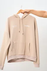 Conjunto deportivo color beige compuesto por un canguro con capucha y un pantalón jogging. El canguro tiene un bolsillo tipo canguro y estampados en el frente. El pantalón es de corte recto con cintura ajustable con cordón.