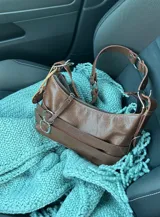 Cartera bandolera marrón de cuero con correa de hombro ajustable, cierre superior con cremallera y detalles de hebillas y aros.