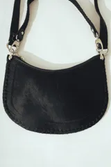 Cartera de hombro negra con forma de media luna, confeccionada en material con textura de pelo o cuero. Presenta costura decorativa gruesa en el borde y correa ajustable con herrajes metálicos plateados.