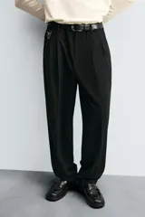 Pantalón gris de corte ancho con cintura elástica, pliegues frontales, bolsillos laterales y traseros de vivo. Incluye detalle de hebilla metálica en la trabilla y cierre frontal con cremallera y botón.