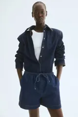 Short de denim azul oscuro, de corte holgado, con cintura elastizada y cordón de ajuste. Cuenta con bolsillos laterales y aberturas laterales en el ruedo.