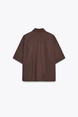 Camisa marrón de popelín con cuello solapa, manga corta con vuelta, bolsillo de plastrón en el pecho, bajo con aberturas laterales, pinzas en la espalda y cierre frontal con botones ocultos.