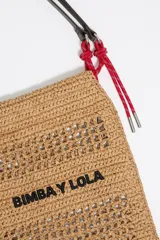 Bolso tote de rafia color beige con diseño calado, asas cortas de cuero marrón y cordones rojos decorativos.