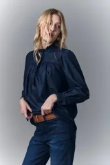 Camisa de denim azul oscuro con cuello mao, corte recto, mangas largas con puños abotonados, cierre frontal con botones a presión y detalle de volados en el cuello y la pechera.