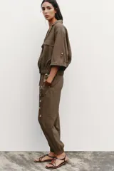 Camisa marrón de cuello solapa y manga corta con trabilla y botón. Tiene bolsillos delanteros de plastrón con solapa, bajo elástico y cierre frontal con botones a presión.