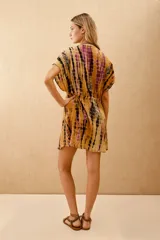 Vestido corto con estampado tie-dye en tonos mostaza, morado y negro. Presenta escote cruzado en V, mangas cortas tipo kimono y cintura ajustada con elástico. Tiene pequeños bordados de cuentas de colores en el escote.