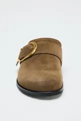 Zueco plano color marrón de serraje, con punta redonda y detalle de tira con hebilla metálica dorada en la parte delantera.