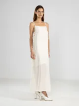 Vestido largo color crudo, de breteles finos y escote recto. Presenta lazos ajustables en los costados, forro hasta arriba de las rodillas y una caída posterior más larga. Confeccionado en tejido arrugado liviano con bolsillos laterales y detalle de Polonio en cobre.