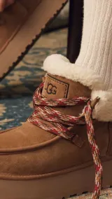 Botines UGG color marrón claro, con interior de piel sintética color crema, cordones de tela con diseño geométrico en marrón, rojo y blanco, y suela de goma color beige.
