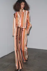 Pantalón de tiro medio con estampado de rayas verticales en tonos marrón, naranja, rosa y beige. Cuenta con trabillas en la cintura, cinturón ajustable con lazadas, bolsillos laterales y cierre frontal con cremallera y botones.
