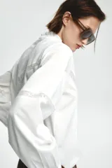 Camisa blanca de cuello alto y manga larga, con detalle de entredós combinados a tono y cierre frontal con botones.